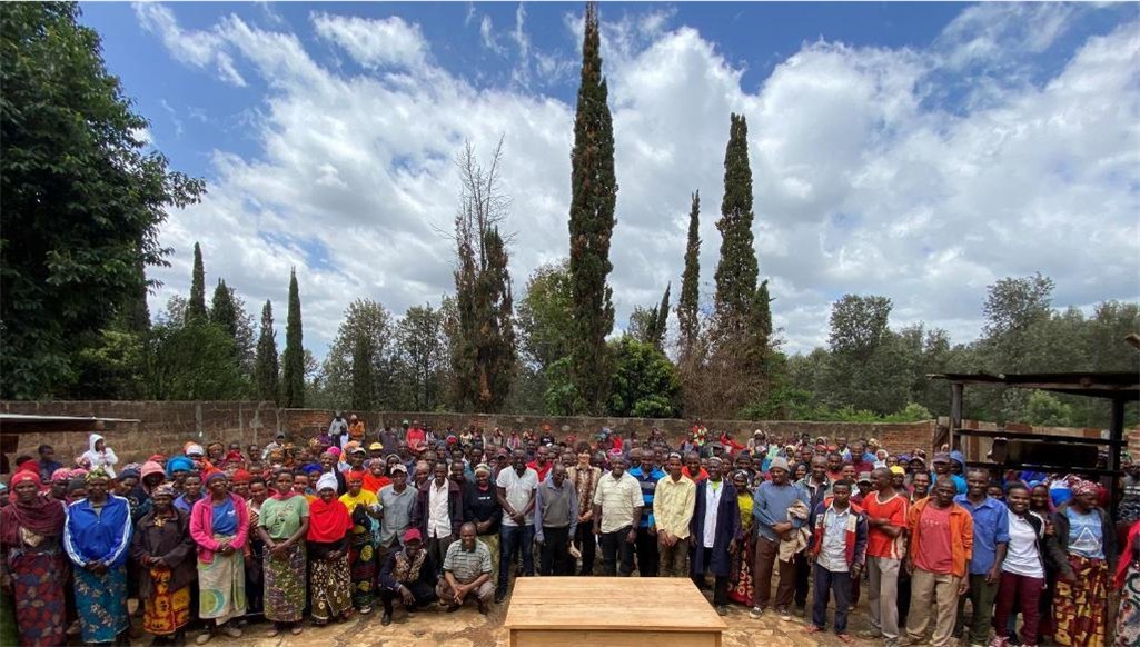 A large group of TANJA workers standing together in front of tall slender trees under a cloudy sky, with a wooden table in the foreground.